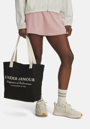 Borsa tote nera in canvas con testo bianco, manici crema, abbinata a una gonna sportiva rosa e sneakers chiare con dettagli arancioni.