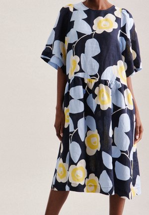 Kobieta w sukience do kolan z krótkim rękawem, z dużym kwiatowym wzorem w kolorach niebieskim, żółtym i białym na czarnym tle.