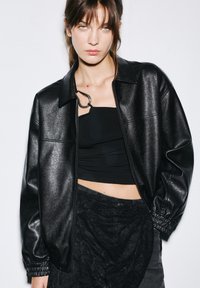 Veste en simili cuir noir avec fermeture éclair, col chemise et poignets élastiques, associée à un haut noir ajusté et une jupe en dentelle superposée.