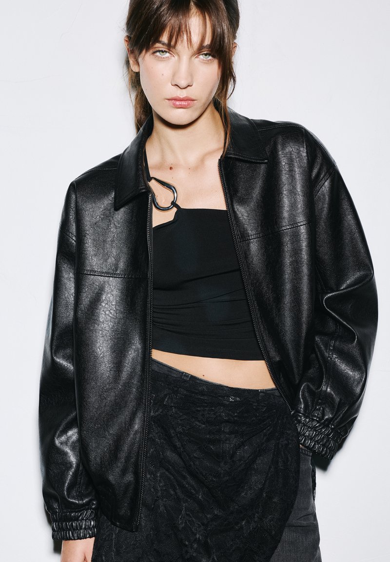 Veste en simili cuir noir avec fermeture éclair, col chemise et poignets élastiques, associée à un haut noir ajusté et une jupe en dentelle superposée.