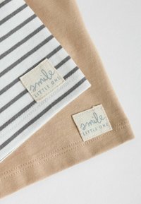 Deux vêtements pour bébé pliés, un beige et un blanc à rayures grises, chacun avec une étiquette en tissu portant l'inscription « smile LITTLE ONE » près de l'ourlet.