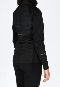 Veste noire avec une partie supérieure matelassée, un col montant et des accents dorés. Comprend des manches longues et une coupe ajustée.