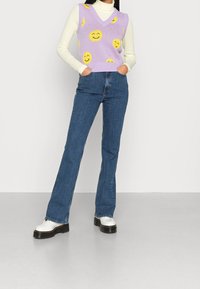 Chaleco de punto morado con patrón de caras sonrientes, superpuesto a un cuello alto blanco. Vaqueros acampanados azules y zapatos de plataforma blancos completan el conjunto.
