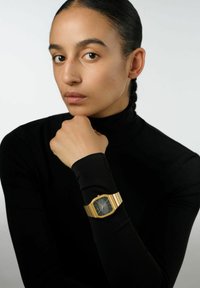 Montre en métal doré avec un cadran noir et des accents argentés, portée sur un poignet avec un haut à manches longues noir, montrant un design octogonal minimaliste.