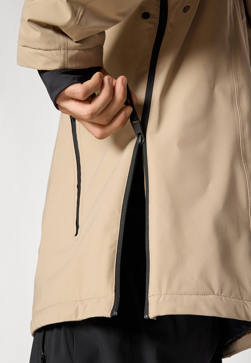 Veste imperméable beige avec fermeture éclair à l'avant et accents noirs. Le tissu a une texture lisse avec un design de couture distinctif.