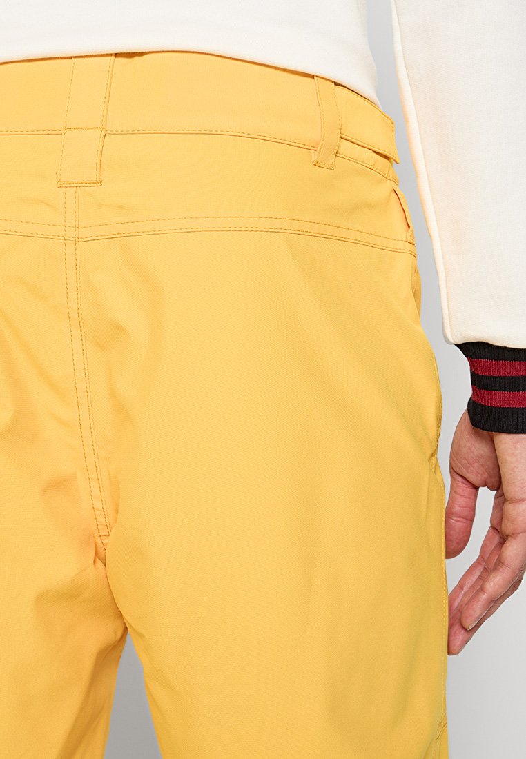 Gelbe Hose aus glattem, strukturiertem Stoff. Mit sichtbaren Nähten, geradem Schnitt und einer Seitentasche, kombiniert mit einem weißen Longsleeve-Oberteil.