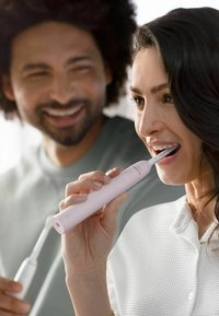 Elektrische tandenborstel met een gladde roze handgreep, slank ontwerp en witte borstelharen; de gebruiker lijkt betrokken terwijl hij zijn tanden poetst.