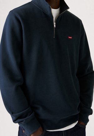 Mann trägt einen marineblauen Sweatshirt mit Viertel-Reißverschluss über einem weißen Hemd, mit Levi's-Logo auf der linken Brust und Händen in den Jeans-Taschen.