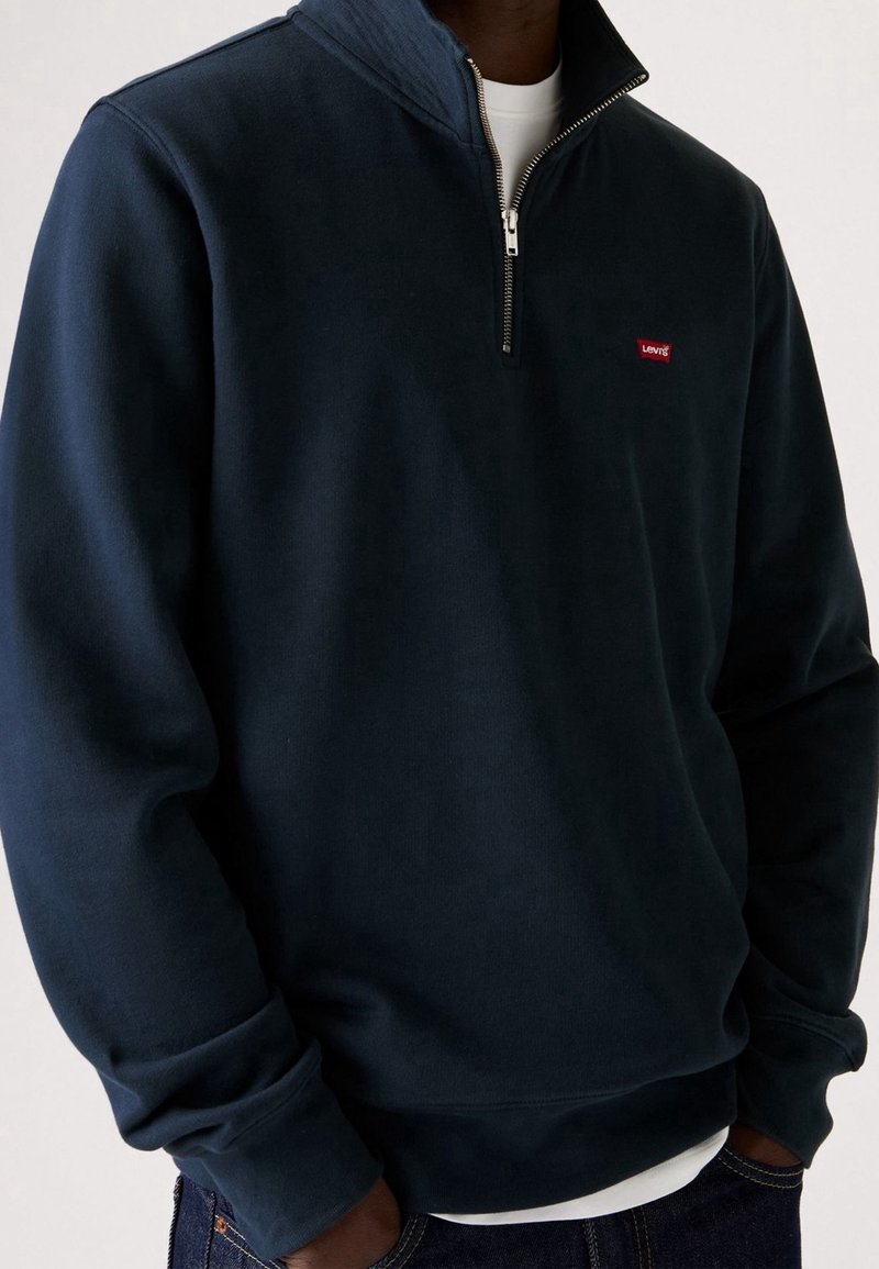 Homme portant un sweat-shirt bleu marine à fermeture éclair sur le quart, superposé sur une chemise blanche, avec le logo Levi's sur la poitrine gauche et les mains dans les poches du jean.