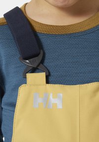 Suspensores amarillos con un acabado texturizado sujetados por un clip de plástico negro, usados sobre una camisa de punto azul con ribete marrón claro. Logo plateado visible.