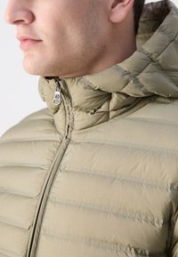 Svetlo olivnozeleni pufast plašč s prešito teksturo, zadrgo in kapuco. Ima ožje vodoravne šive za izolacijo.