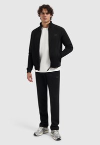 Zwarte zip-up jack met hoge kraag en zijzakken, gecombineerd met een witte t-shirt en zwarte joggers. Grijze sportschoenen maken de outfit compleet.