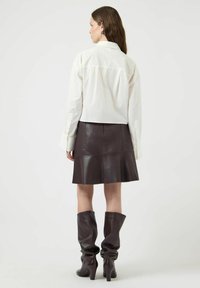 Wit overhemd met knopen, bruine leren rok die uitloopt bij de zoom, kniehoge bruine laarzen met een nonchalante textuur en puntige hakken.