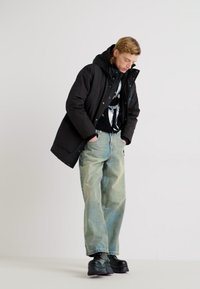 Cappotto imbottito nero, vestibilità oversize, con cappuccio grande. Sotto, un maglione grafico nero, jeans ampi blu chiaro e stivali neri chunky.