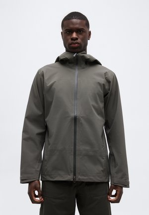 Egy férfi szürke kapucnis, vízálló dzsekit visel, amelynek elején cipzár található, és egy egyszínű fehér háttér előtt áll.