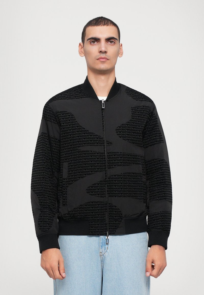 Black bomber jacket with textured logo pattern, zip closure, and ribbed cuffs. Smooth fabric, casual design, and relaxed fit.