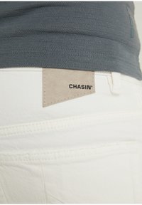 Vaqueros de denim blanco con una etiqueta de cuero beige que muestra "CHASIN'" cosida en la parte trasera. Parte de una camiseta azul texturizada es visible por encima de la cintura.