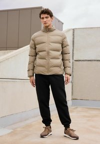 Jachetă puffer bej cu guler înalt, fermoar complet pe față, pantaloni de trening negri și adidași căprui. Material neted, design izolat, manșete ajustate.