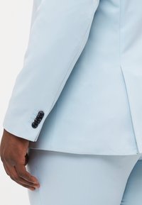 Helle blaue Sakko mit glattem Stoff, strukturiertem Design und taillierter Passform. Schwarze Knöpfe und dezente Stickdetails an den Ärmeln.