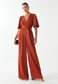 Mono de color óxido con escote en v profundo y amplias mangas acampanadas. Fabricado en un tejido suave; presenta una cintura ajustada y un corte de pierna amplia.