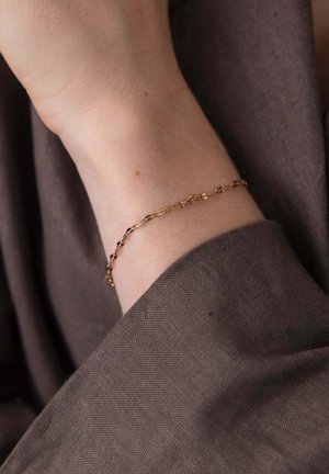Delicate gold chain bracelet on a wrist, resting against soft brown fabric background.