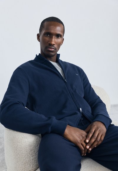 Marineblå zip-up jakke med høj krave, ribbede ærmer og et diskret logo på brystet, lavet af et blødt, struktureret stof.