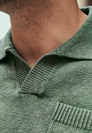 Close-up van iemands nek en kaak met lichte stoppels, die een gestructureerd groen gebreid poloshirt draagt met een opgevouwen kraag.