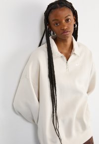 Ανοιχτό μπεζ φούτερ με φερμουάρ και ψηλό γιακά, σε oversized εφαρμογή, με ασημένιο λογότυπο της Nike. Υφασμάτινη υφή με χαμηλωμένα ώμους.