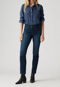 Kvinna som bär en mörkblå denimskjorta med uppvikta ärmar, mörka skinny jeans och svarta klackstövletter, stående mot en enhetlig bakgrund.