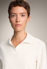 Pulover alb cu guler, cu o textură cu nervuri și detaliu în formă de V. Modelul are părul scurt și un ton al pielii neutru, fiind încadrat într-un fundal cu un gradient delicat.