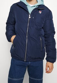 Mörkblå puffjakka med dragkedja framtill, två sidofickor och en bröstficka. Har elastiska ärmslut och dragsko i fållen. Strukturerad yta.