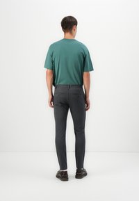 Camiseta de algodón verde, de manga corta, combinada con pantalones gris ajustados que cuentan con dos bolsillos traseros y un patrón sutil. Unos zapatos marrones completan el look.
