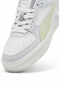 Fehér és világosszürke sneaker perforált orral, texturált bőr és szintetikus díszítésekkel, lapos fehér fűzőkkel és gumitalppal.