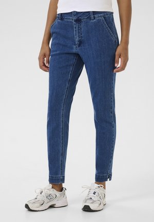 Person wearing blue denim jeans and white sneakers, standing against a plain white background.