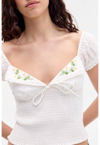 Top bianco arricciato con maniche corte a palloncino, scollatura a v con pizzo, ricamo floreale verde con tocchi di giallo. Dettaglio a fiocco nella parte anteriore.