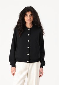 Black cardigan with a herringbone texture, round neckline, and shiny silver buttons; features two side pockets and ribbed cuffs.