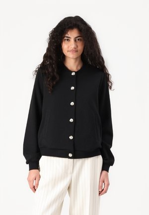 Cardigan noir avec une texture en chevrons, col rond et boutons en métal argenté brillants ; il possède deux poches latérales et des poignets à côtes.
