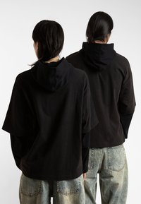 Svart hettegenser med design i to lag, som har en ensfarget svart utside og et teksturert innersjikt. Oversized passform med vide ermer.
