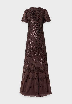 Mujer de pie vestida con un vestido largo hasta el suelo de color marrón oscuro con mangas cortas y patrones florales con lentejuelas negras, combinado con tacones abiertos de color claro.