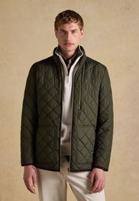 Mann trägt eine olivgrüne gesteppte Jacke über einem weißen Pullover mit Viertelreißverschluss und beigen Hosen, steht vor einem einfarbigen beigen Hintergrund.