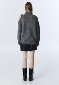 Maglione grigio a maglia con collo alto, vestibilità over, e orlo sfilacciato, abbinato a una gonna plissettata nera e stivaletti neri lucidi.