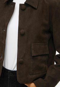 Brun ruskindsjakke med krave, frontknapper og en patch-lomme. Materialet har en blød tekstur; den er cropped og tætsiddende ved taljen.