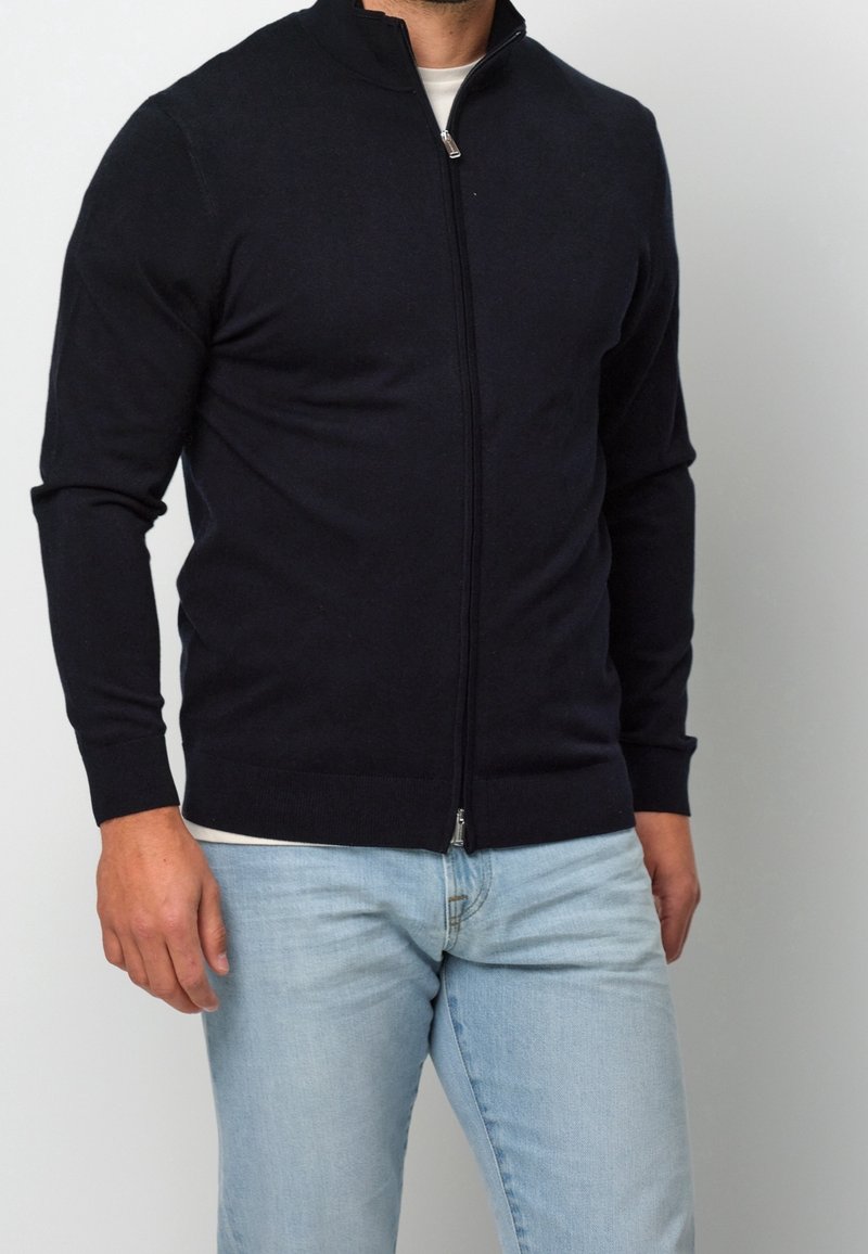 Homme portant un pull noir à fermeture éclair sur une chemise blanche et un jean bleu clair, sur un fond uni clair.
