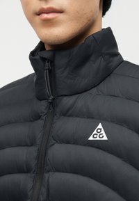 Κοντινό πλάνο ενός ατόμου που φοράει μαύρο μπουφάν puffer με ψηλό γιακά και λευκό κεντημένο λογότυπο ACG στο στήθος.