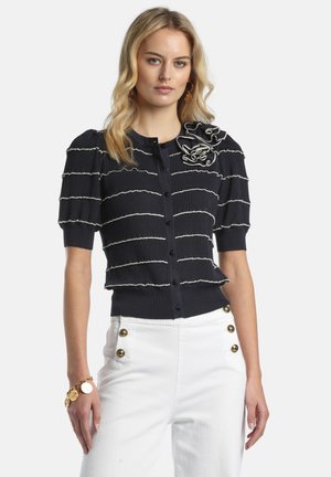 Vrouw draagt een zwart kort mouwig, geknoopt shirt met witte horizontale strepen en witte broek met gouden knopen, poserend met een gouden armband.