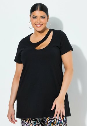 Lächelnde Frau mit glattem Haar trägt ein schwarzes Kurzarmoberteil mit asymmetrischem Ausschnitt und bunte Zebra-Print-Hose, steht vor einer weißen Wand.