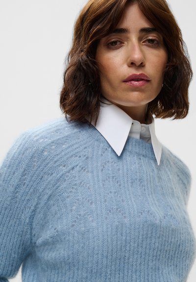 Femme aux cheveux bruns mi-longs portant un pull en tricot bleu clair par-dessus une chemise blanche à col, sur un fond uni.