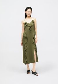 Mulher em pé, vestindo um vestido midi sem mangas em verde-oliva com babados e fenda, combinado com sapatos de salto em cunha pretos e de bico aberto, em fundo plain.