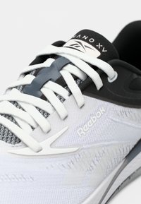 Białe sportowe buty z teksturowanym siateczkowym cholewką, szarymi akcentami i białymi sznurowadłami. Posiadają wyraźne logo Reebok i gładki kształt podeszwy.