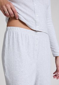Světlé šedé domácí oblečení zahrnující top na knoflíky a kalhoty s elastickým pasem. Obsahuje žebrovanou strukturu a uzavírání kalhot na pět knoflíků.